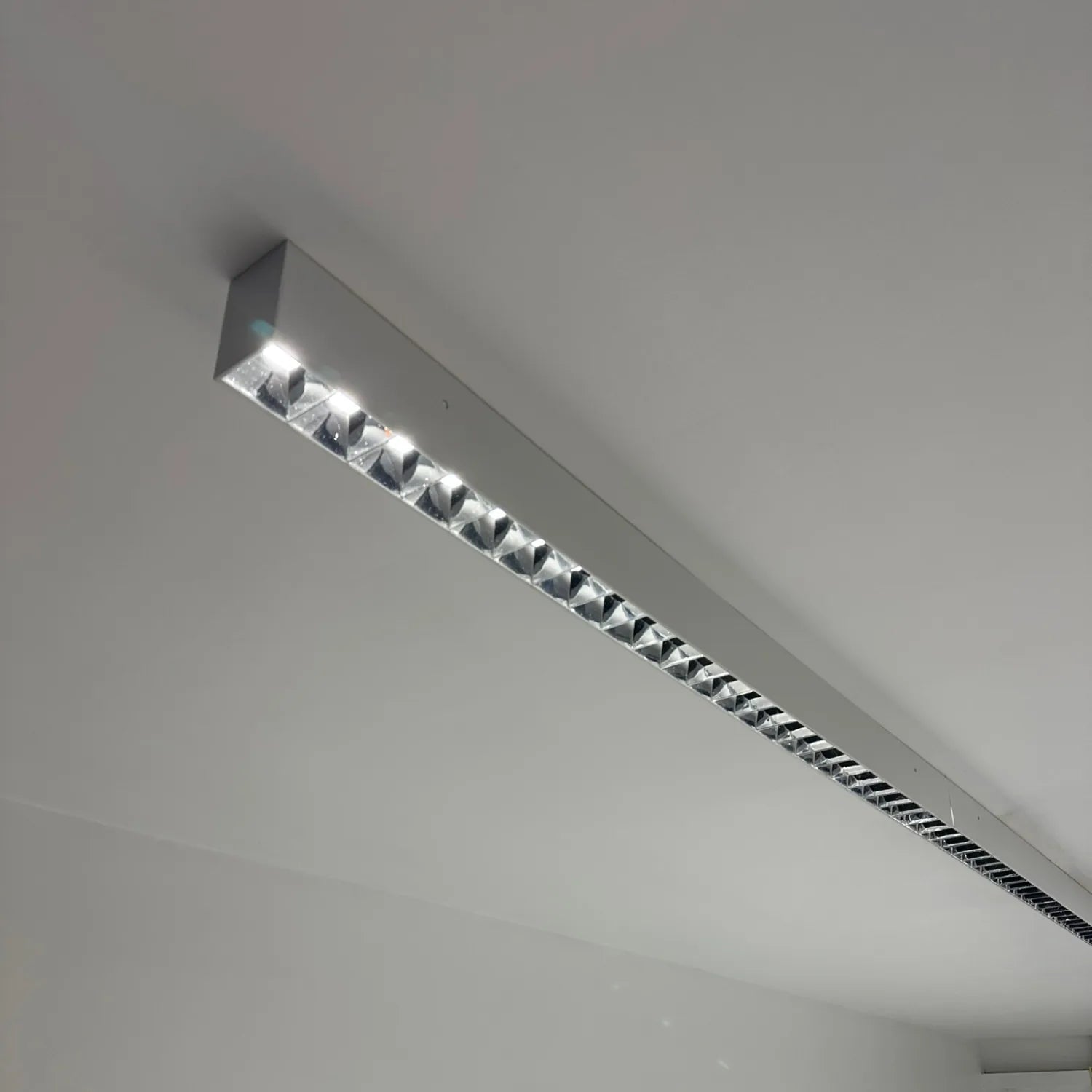 Linear LED batten light mounted on white ceiling, showing aluminum housing with integrated downlights along its length.