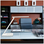 White LED desk lamp with wireless charger base on modern glass desk in office setting with blue chair and storage shelving be