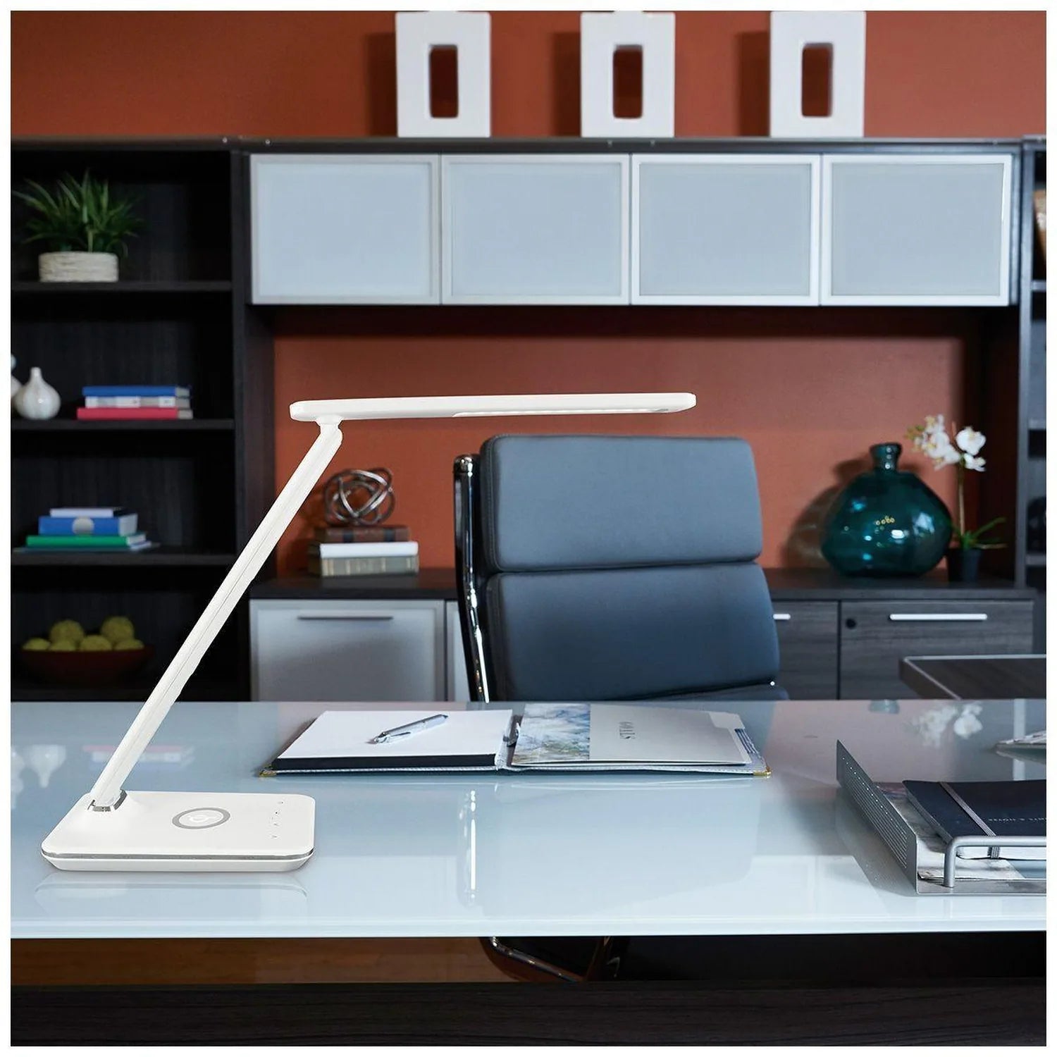 White LED desk lamp with wireless charger base on modern glass desk in office setting with blue chair and storage shelving be
