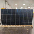 Large black bifacial solar panel on wooden pallet displayed indoors under fluorescent lights, worker visible in background.