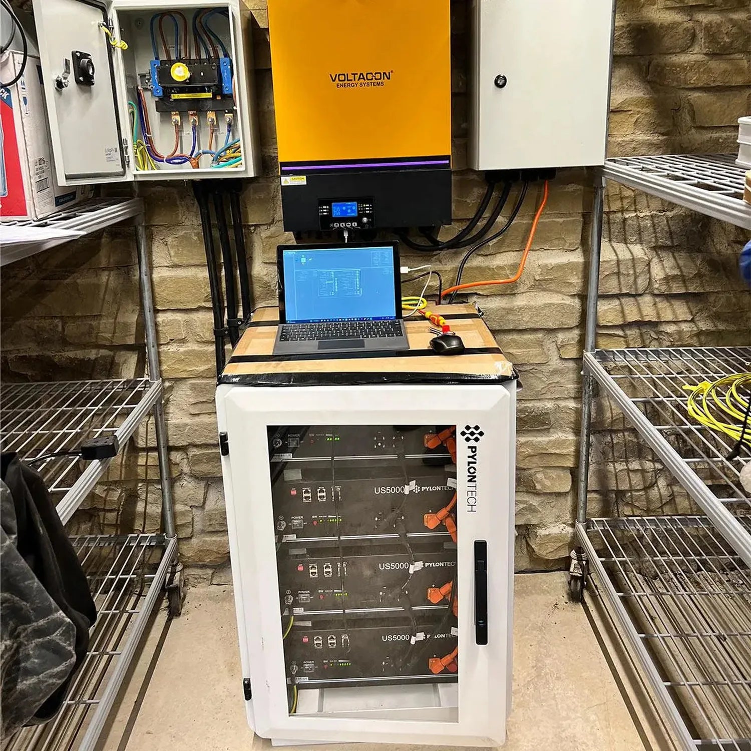 Pylontech US5000 battery cabinet installed in home energy system with Voltacon inverter and monitoring laptop on stone wall.