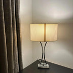 Modern table lamp with curved black metal base and pleated cream cylindrical shade, lit and displayed on dark surface beside 