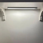 LED under-cabinet light installed on white wall with two white mounting brackets and linear light bar above.
