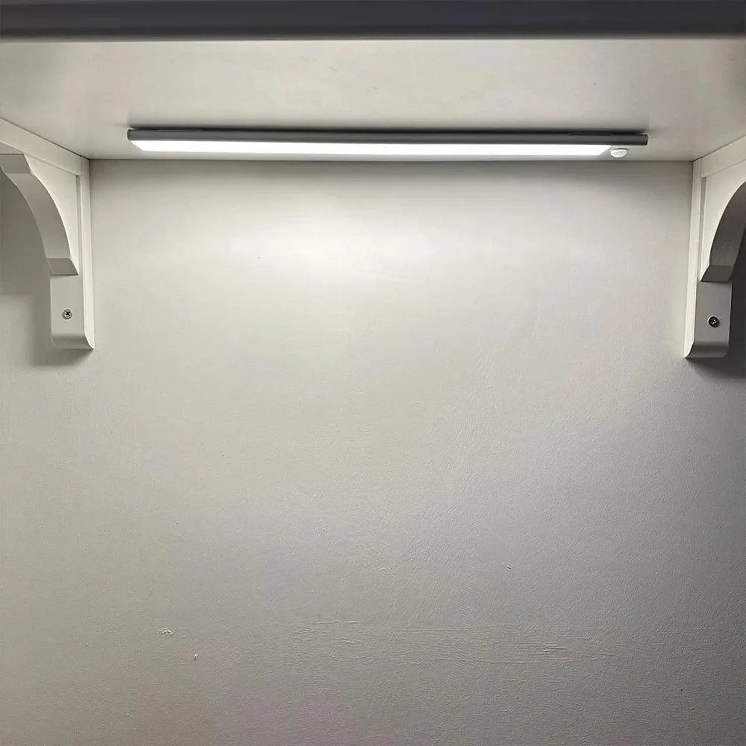 LED under-cabinet light mounted on white wall above empty shelf, with linear fixture and side brackets.