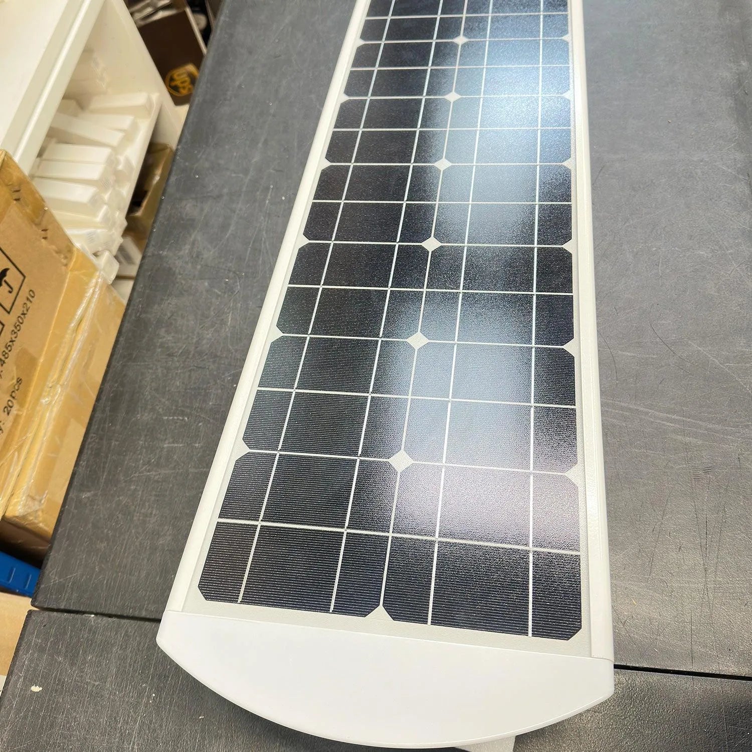 White solar panel with black photovoltaic cells arranged in grid pattern, displayed on concrete surface next to cardboard pac