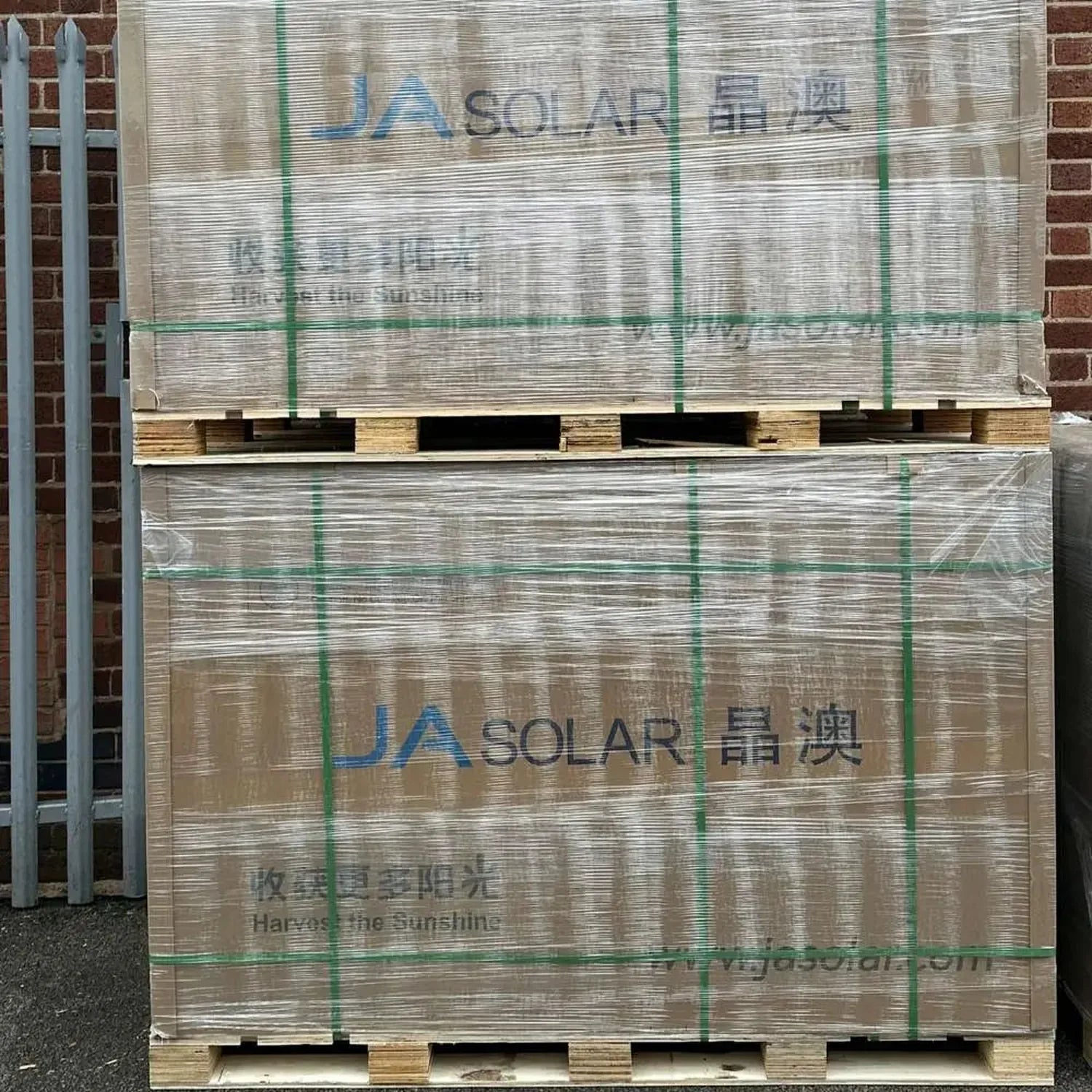 Stacked pallets of JA Solar panels wrapped in plastic with green strapping tape against a brick wall.