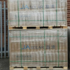 Stacked pallet of JA Solar photovoltaic panels wrapped in plastic with green strapping tape against brick wall.