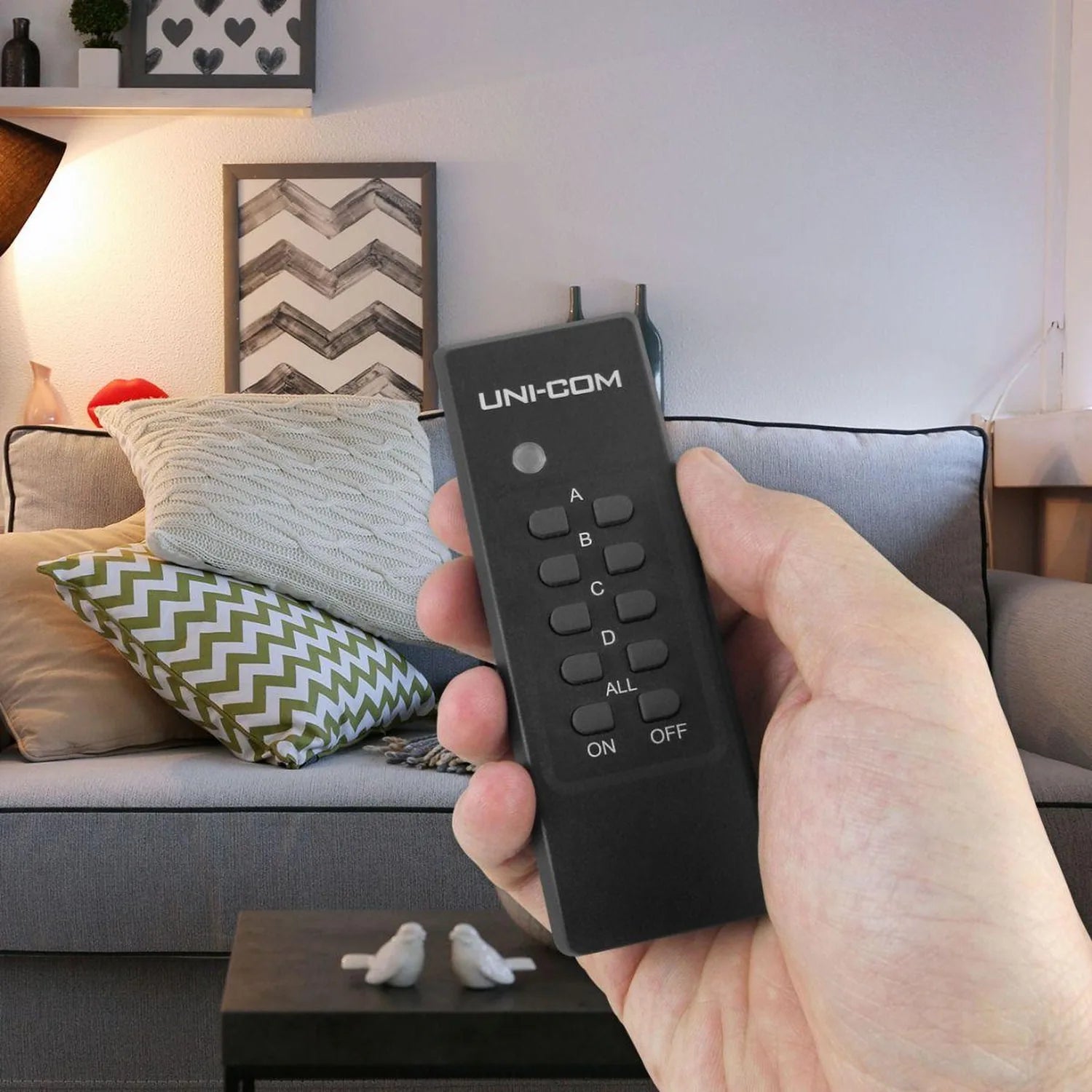 Hand holding black UNI-COM remote control with antenna, displaying buttons labeled A, B, C, D, ALL, ON, OFF, in living room s