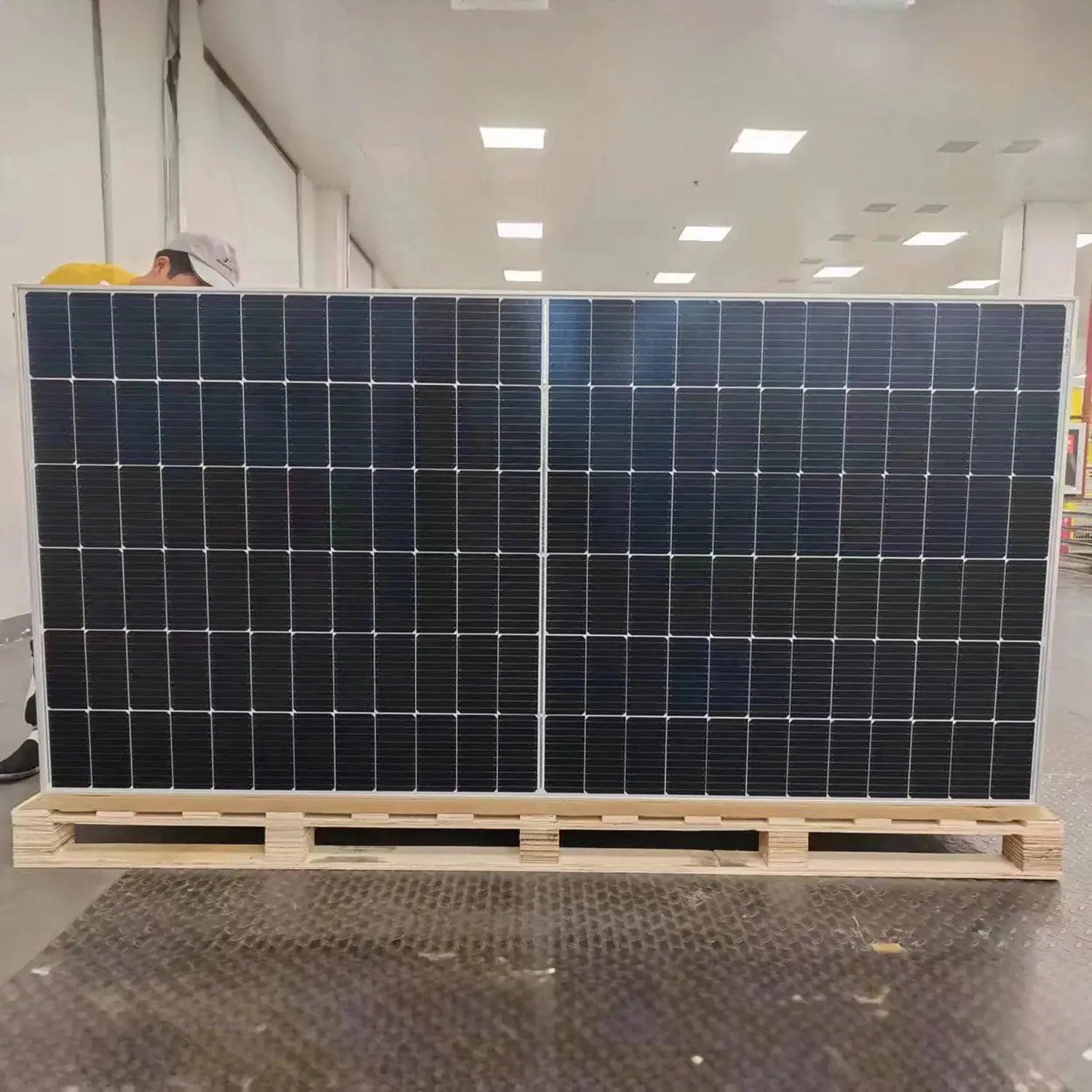 Large bifacial solar panel mounted on wooden pallet in warehouse setting with worker visible in background.