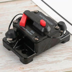 Black waterproof circuit breaker with red manual reset switch and lever, mounted on wooden surface with cable attached.