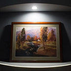 Framed landscape painting with trees and stream displayed on white wall shelf with recessed LED spotlight overhead