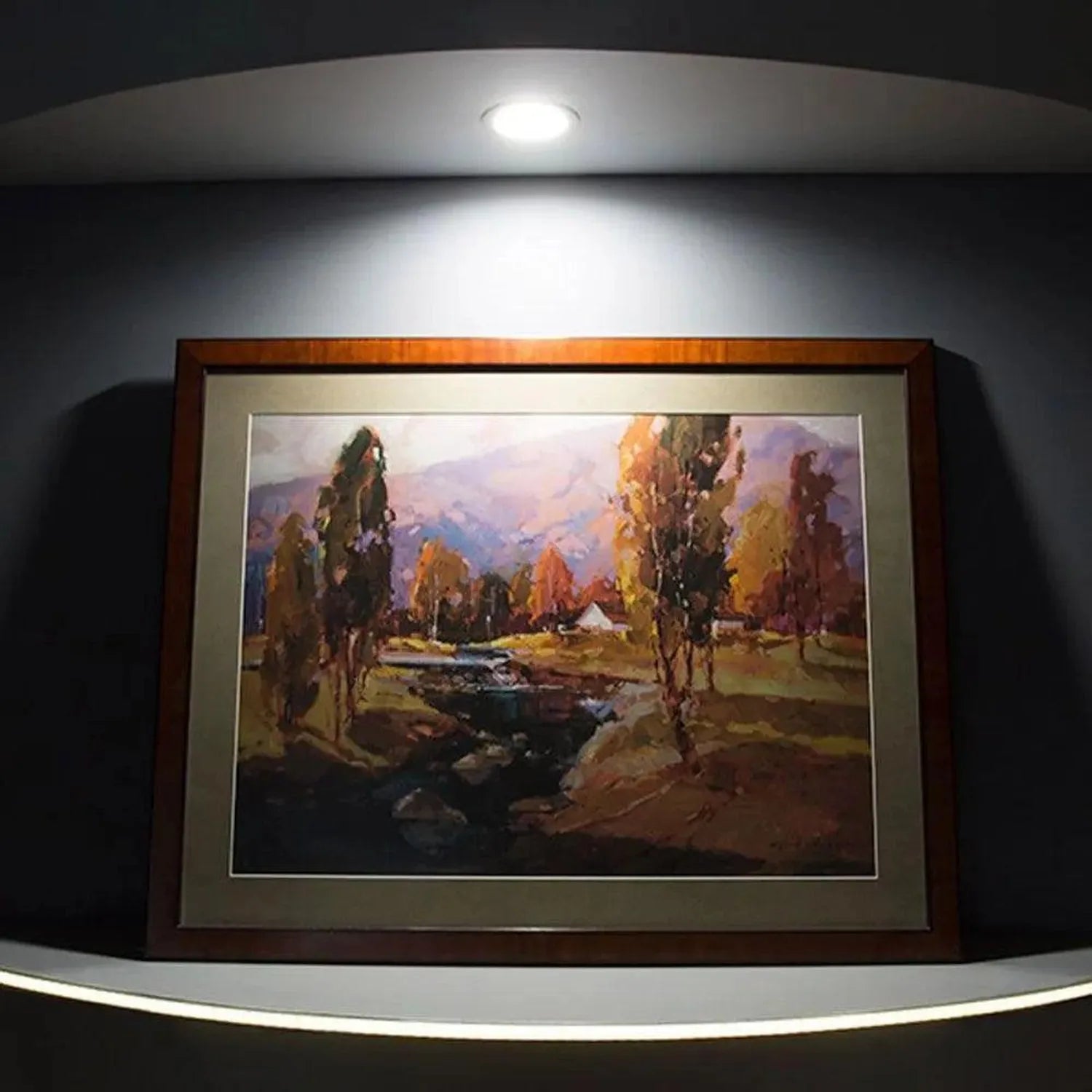 Framed landscape painting with trees and stream displayed on white wall shelf with recessed LED spotlight overhead