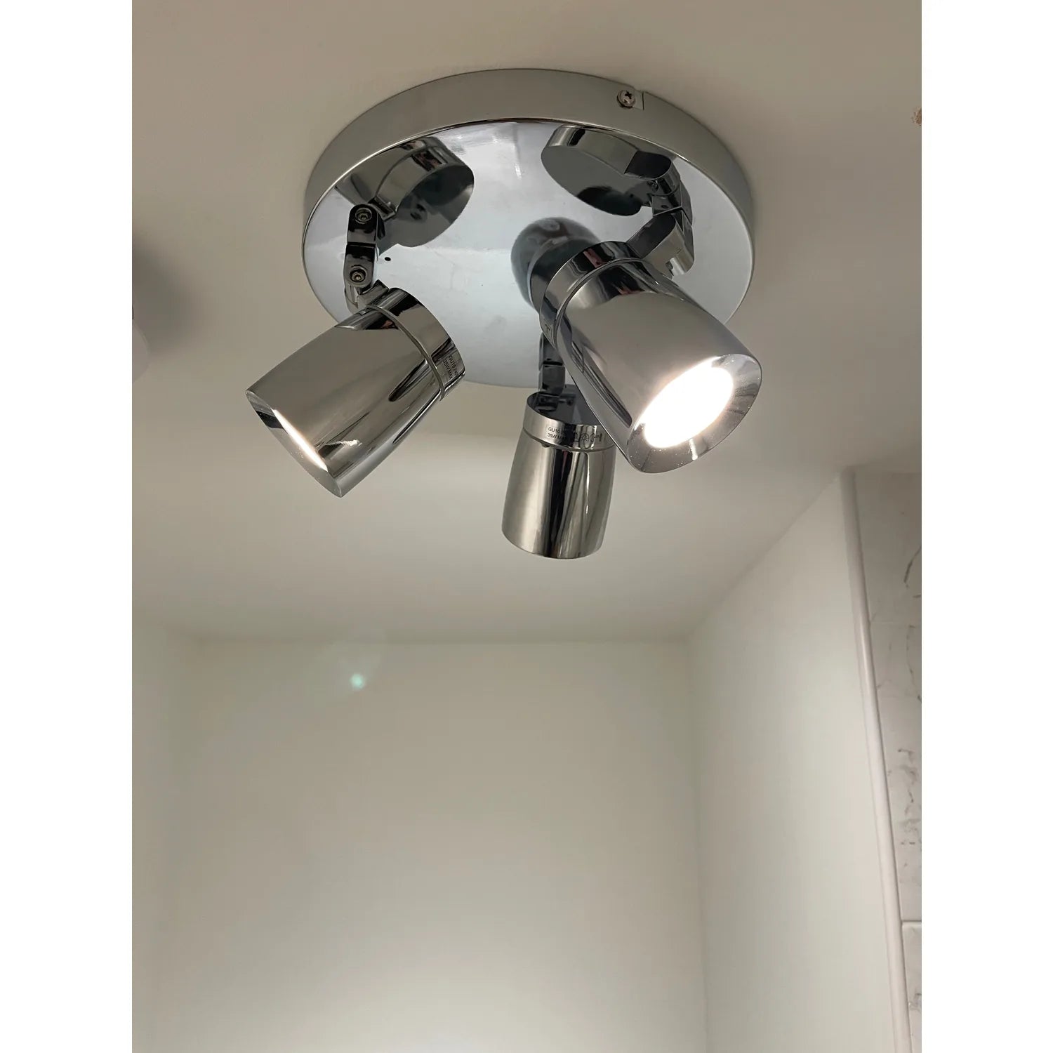 Chrome ceiling spotlight fixture with three adjustable cylindrical spotlights mounted on white ceiling, one bulb illuminated.