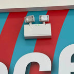 White emergency light unit with dual halogen heads mounted on colorful striped wall, showing installation example.