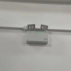 White ceiling-mounted emergency light unit with twin LED spotlights and motion sensor, installed against white ceiling.