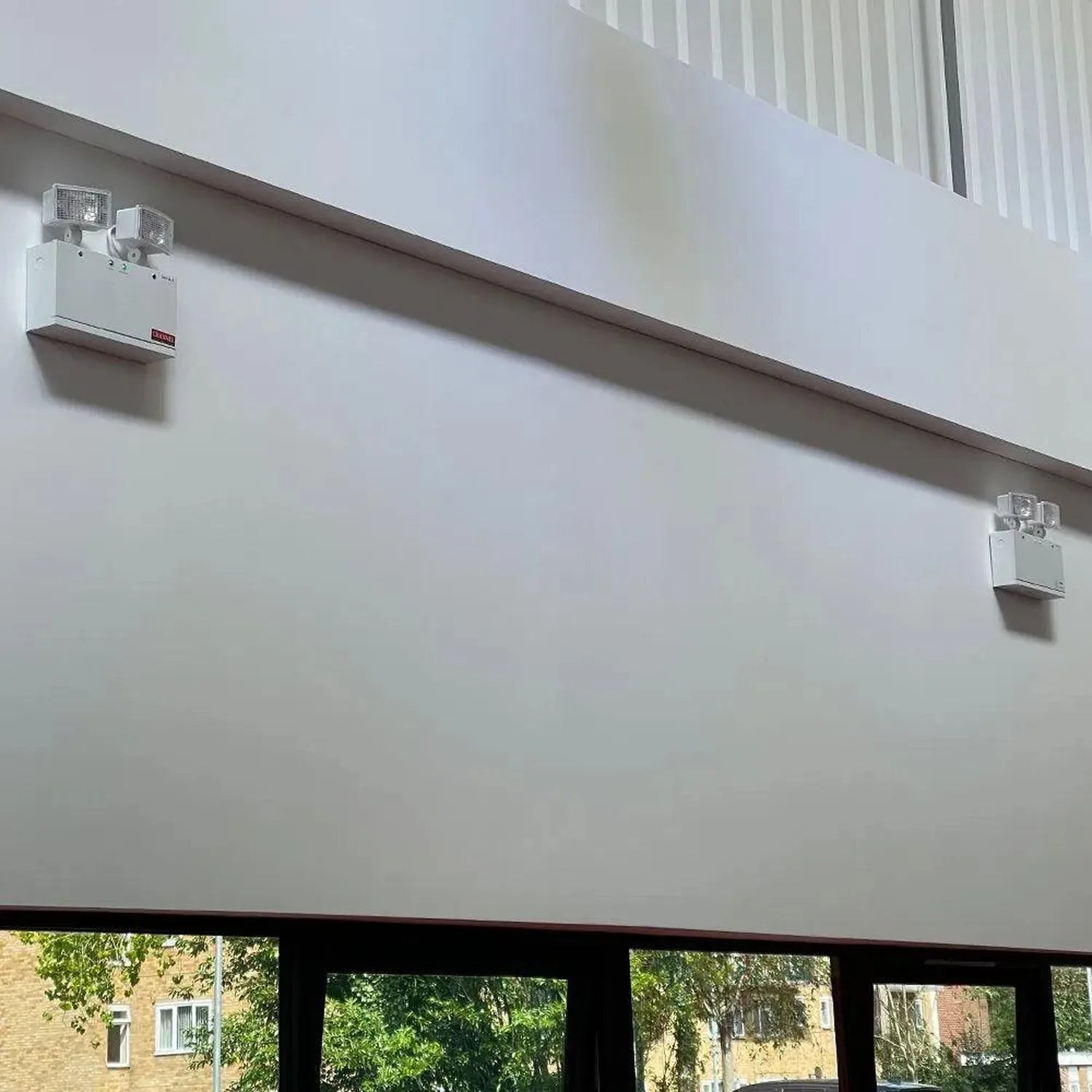 Twin spot emergency LED light mounted on white wall above windows, showing installation on building exterior.