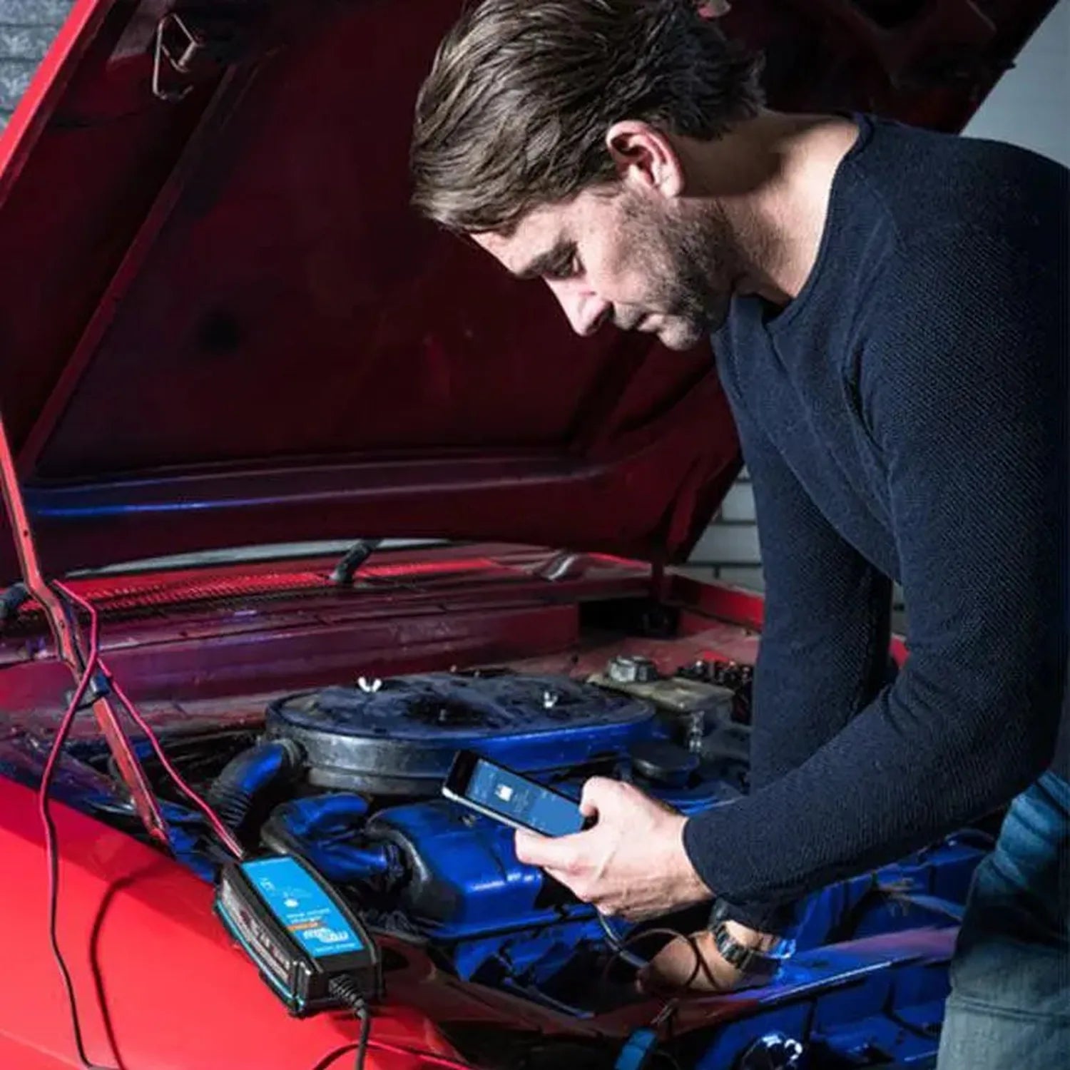 Man using blue Victron charger on red car engine with hood open, demonstrating installation.