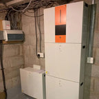 All-in-one hybrid inverter and lithium battery system installed on basement wall, with orange and white casing, electrical wi