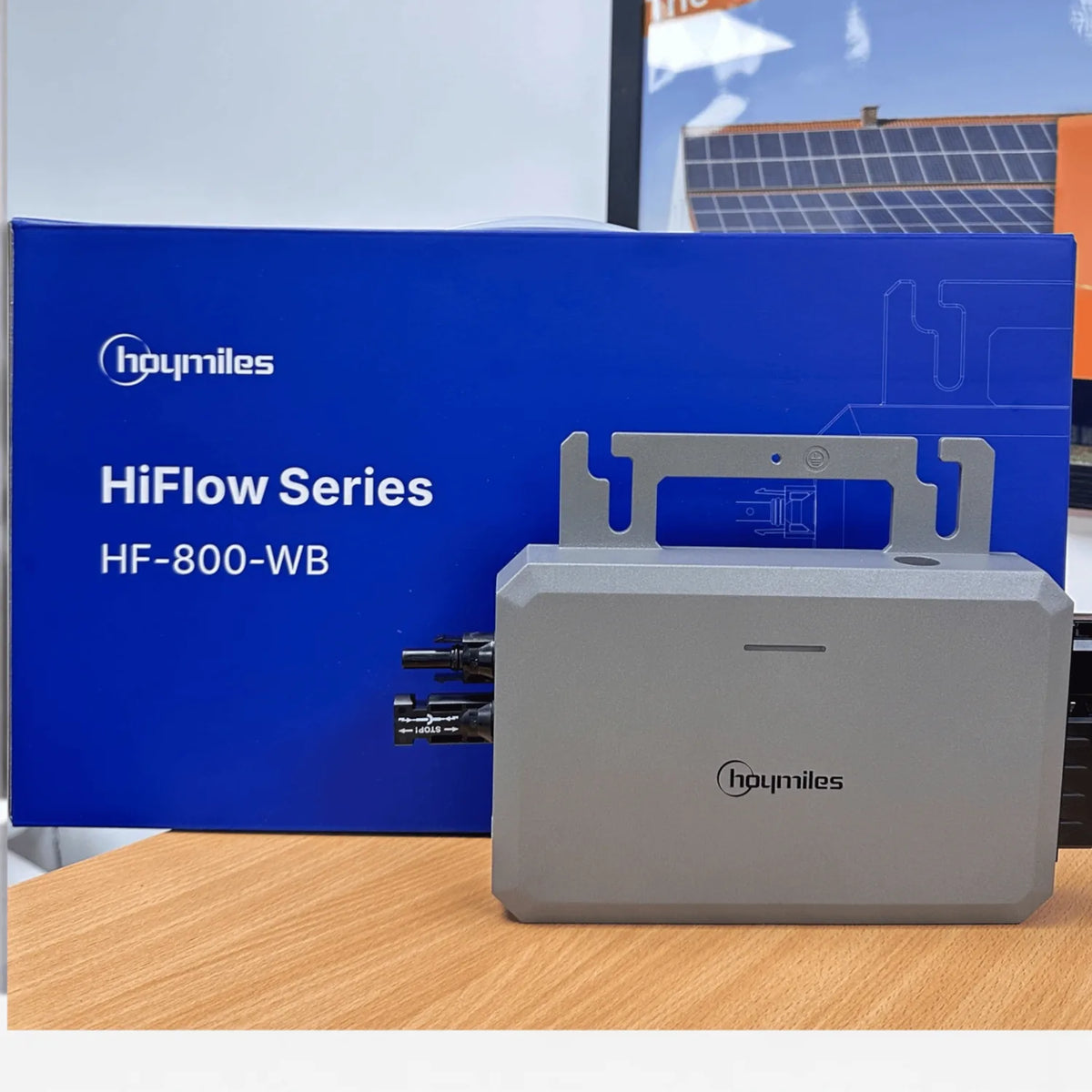 Hoymiles HiFlow Series HF-800-WB micro inverter next to its blue packaging box, displayed on wood with solar panels visible i