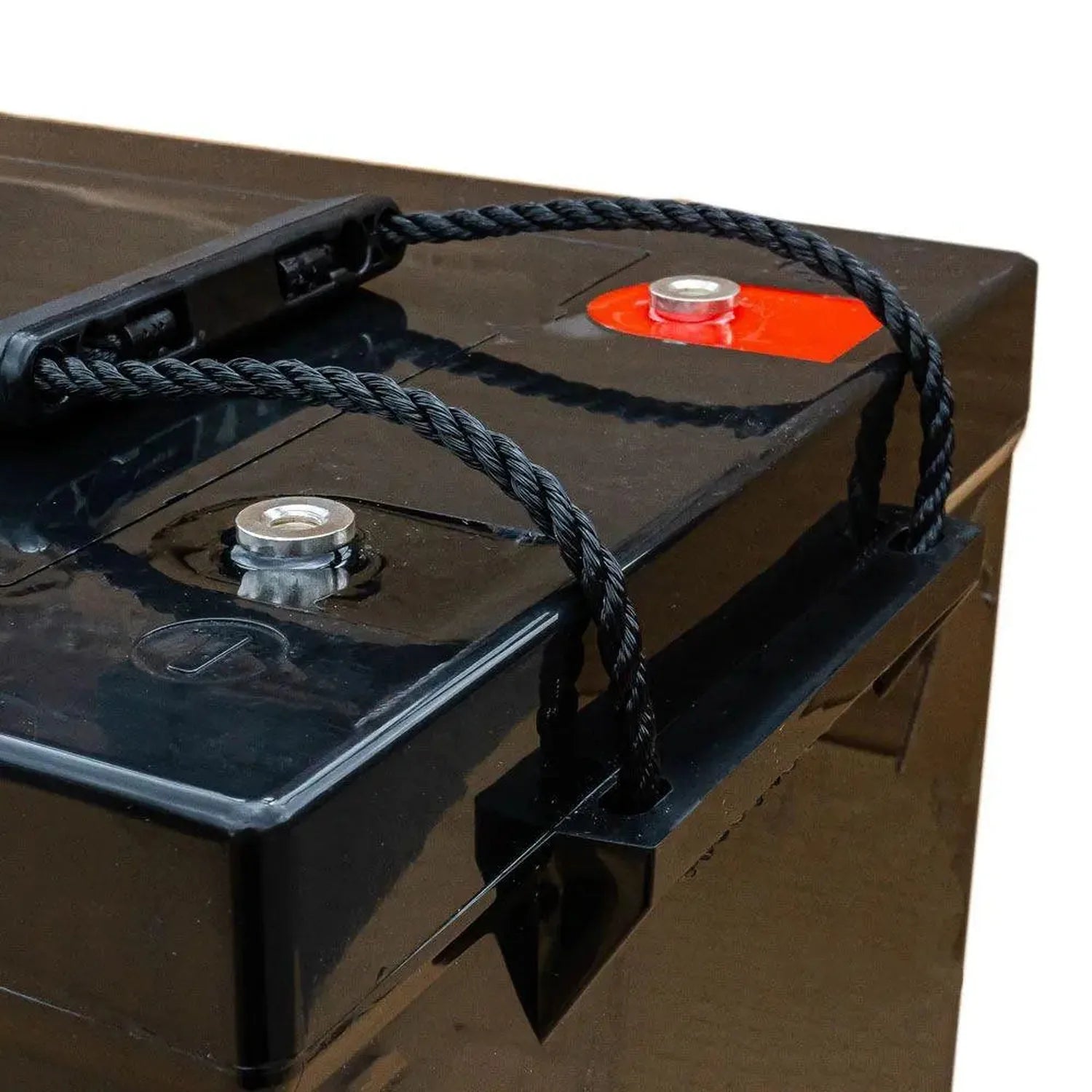 Black deep cycle battery with red terminal cap, mounted on brown surface with black rope handles and metal mounting brackets.