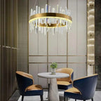 Gold ring pendant light with white crystal rods hanging above round marble table and two navy blue chairs in modern interior 