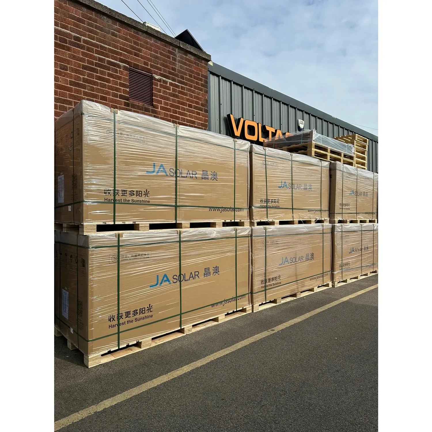 Stacked wooden pallets of JA Solar panels wrapped in plastic, loaded outside a warehouse with VOLTA signage visible above.