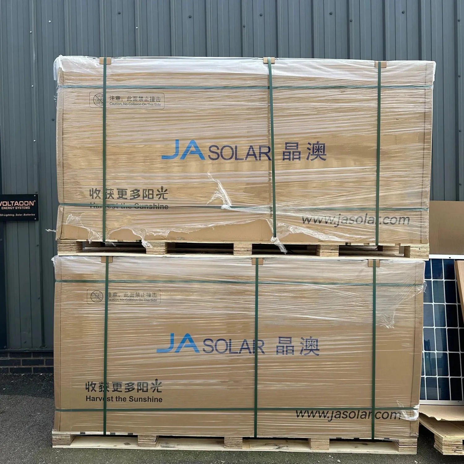 Two large wooden pallets of JA Solar bifacial solar panels stacked against a metal warehouse wall, wrapped in plastic with gr