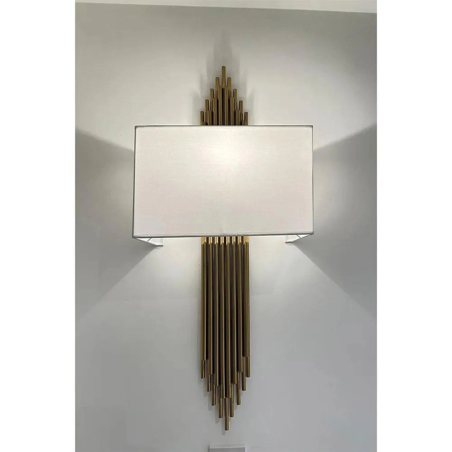 Brass wall sconce with rectangular linen shade and cascading vertical rods mounted on white wall, installation shot showing d