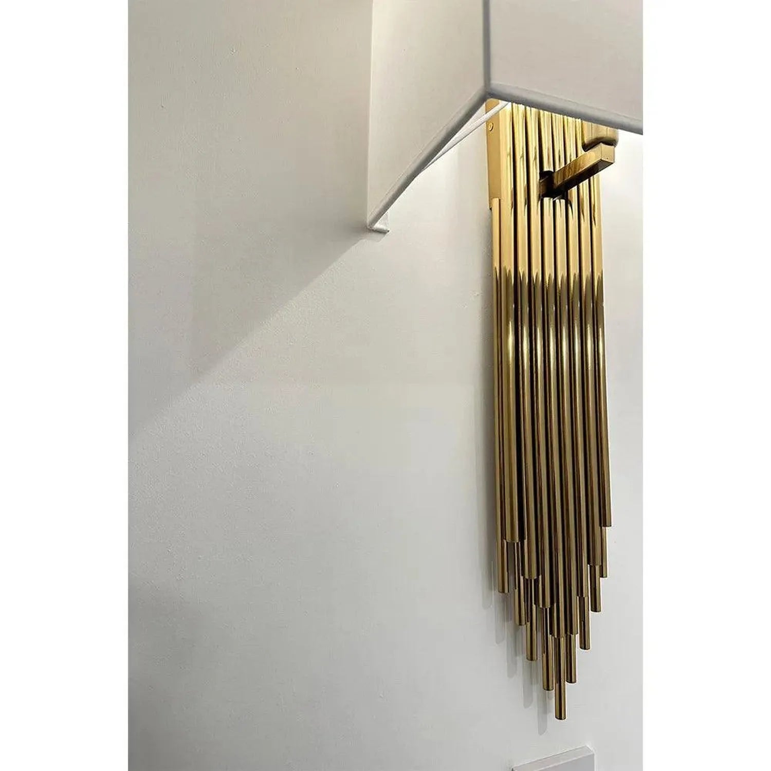 Gold fringe wall sconce with cascading metal rods mounted on white wall with white bracket arm.