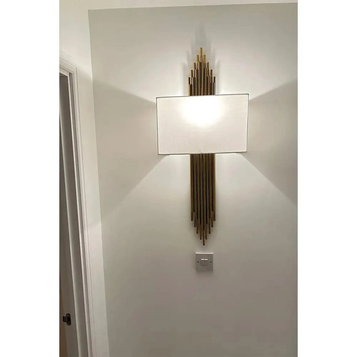Gold vertical lined wall light with rectangular white shade mounted on white wall in modern interior setting.