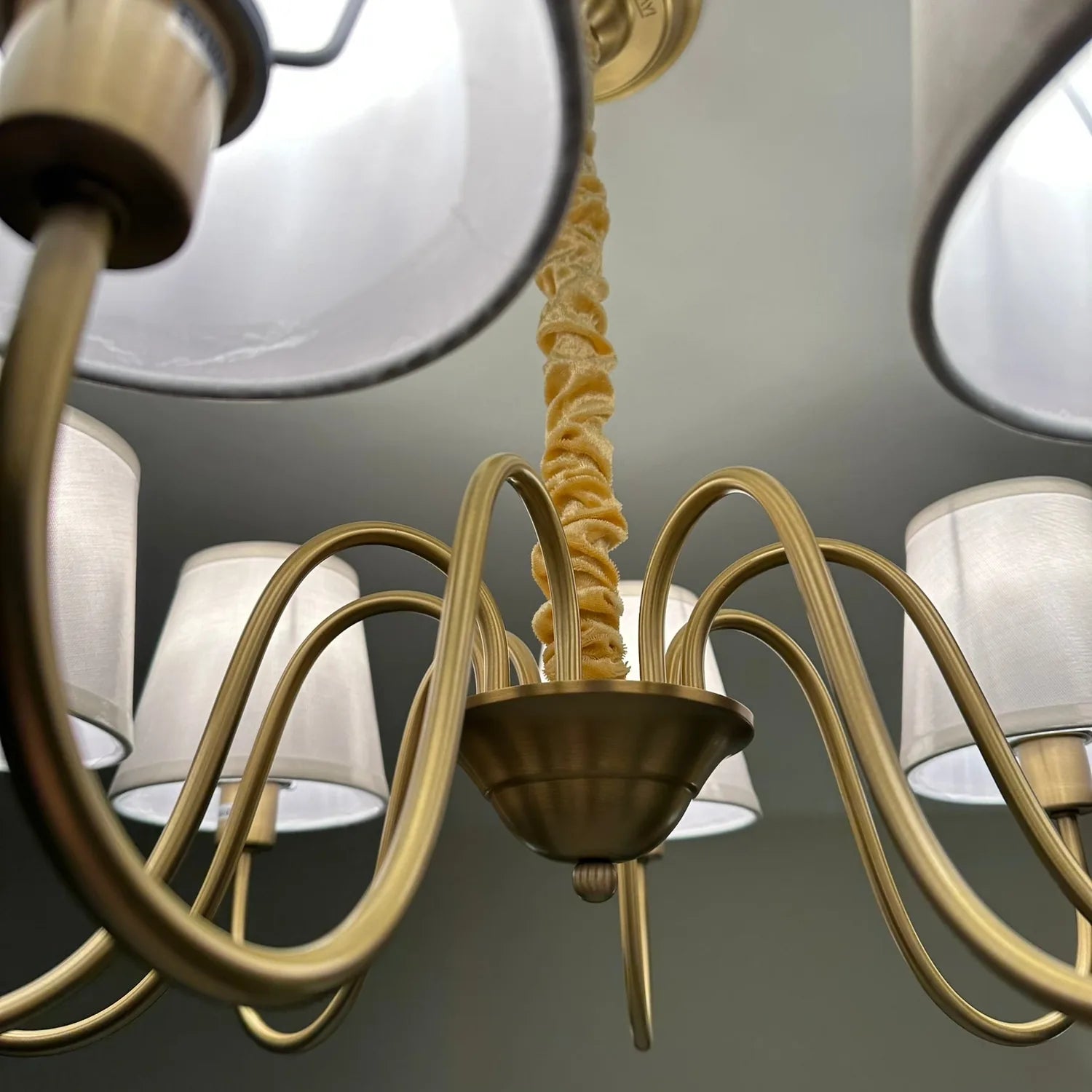 Gold chandelier with eight curved arms and white fabric shades, shown installed against ceiling with decorative rope detail.