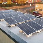 Solar panels installed on flat roof of brick building with concrete mounting blocks, residential area visible in background