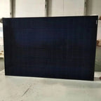 Black monocrystalline solar panel displayed on concrete floor indoors, showing grid pattern of cells.