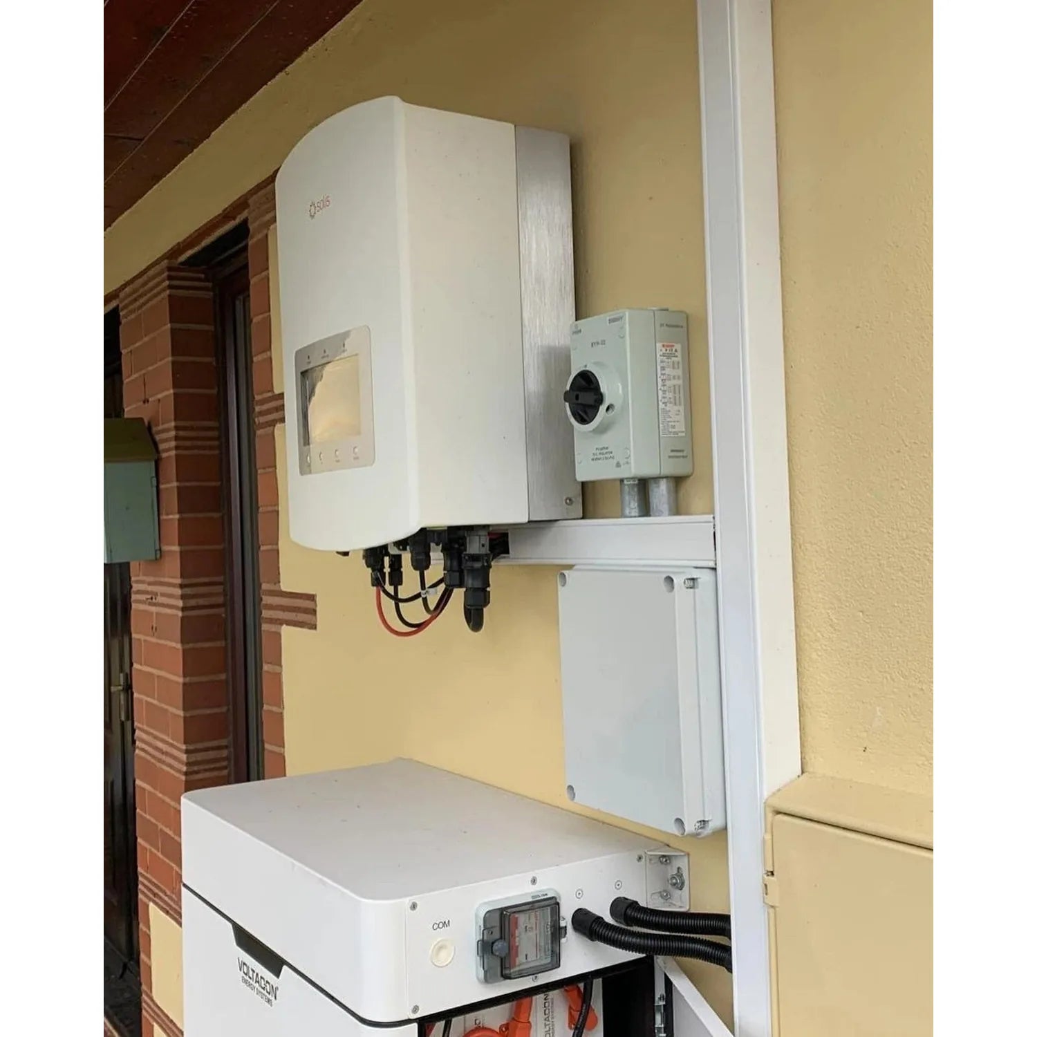 Solis hybrid solar inverter mounted on yellow wall above white battery storage unit in residential installation.