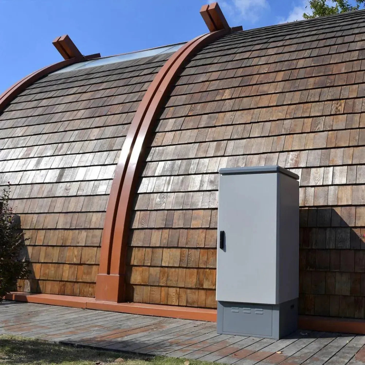 White modular battery enclosure cabinet installed on wooden deck beside curved wooden-sided building with metal accents.