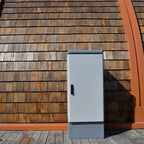 White metal battery enclosure mounted on wooden shingle exterior wall next to orange trim, installation shot.