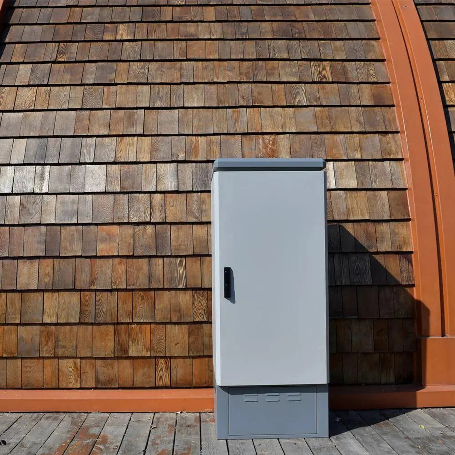 White metal battery enclosure mounted on wooden shingle exterior wall next to orange trim, installation shot.