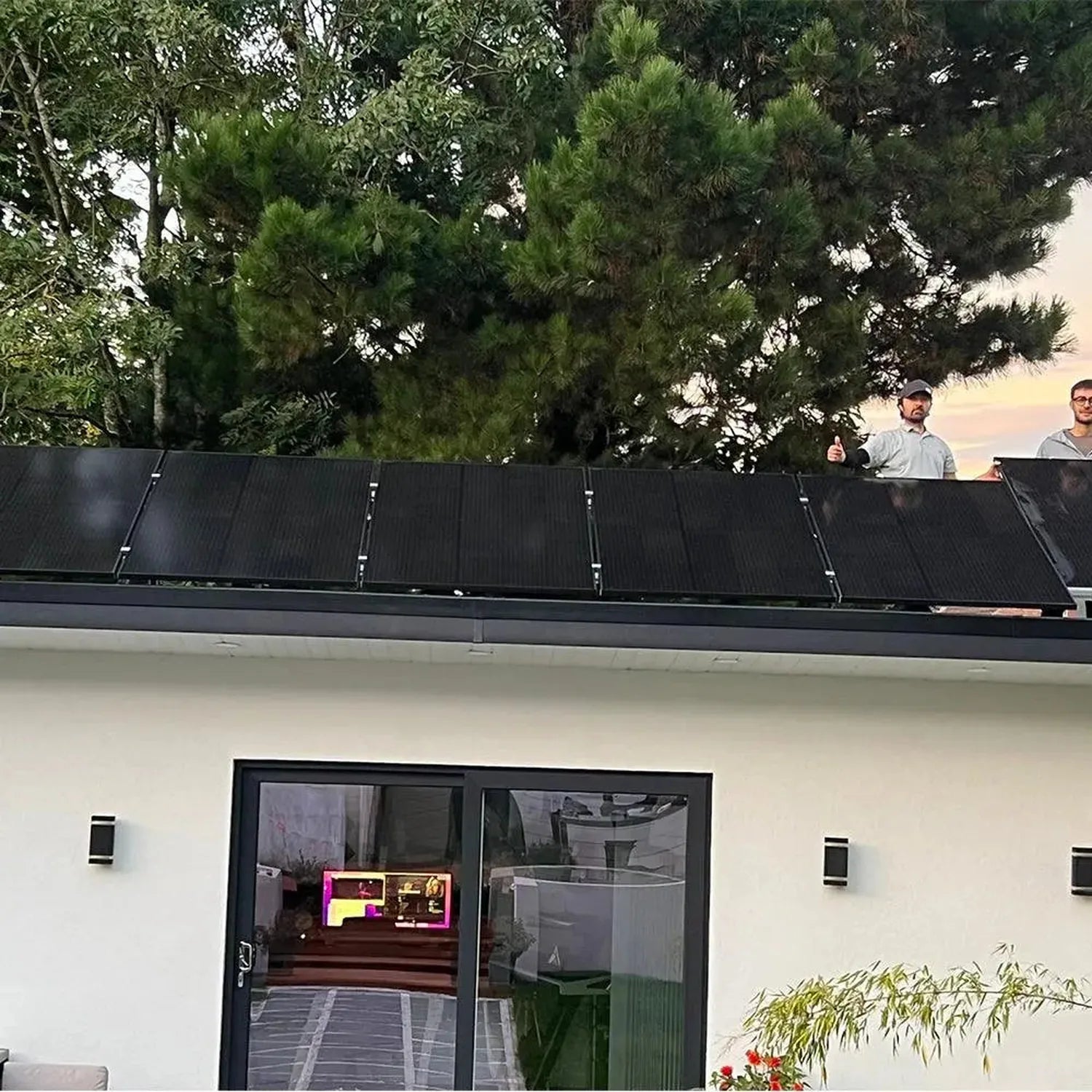 Black solar panels installed on modern white house roof with trees and two people visible above