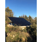 Large black solar panel array mounted on ground in rural landscape with evergreen trees and clear sky.