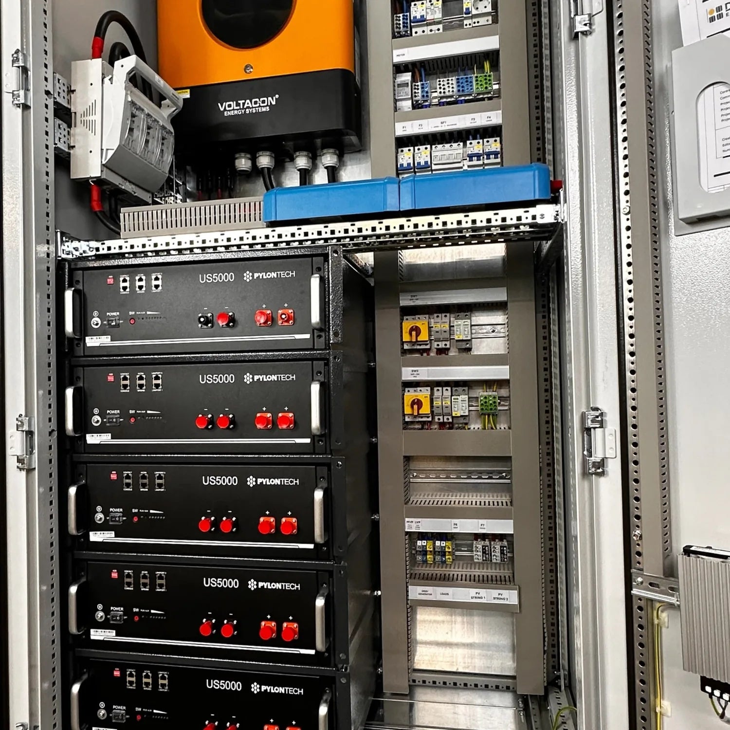 Industrial installation shot showing five stacked Pylontech US5000 battery units with red controls in a metal enclosure, alon