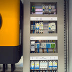 Open electrical cabinet showing circuit breakers, voltage arresters, protection modules, meter, and terminal blocks with blue